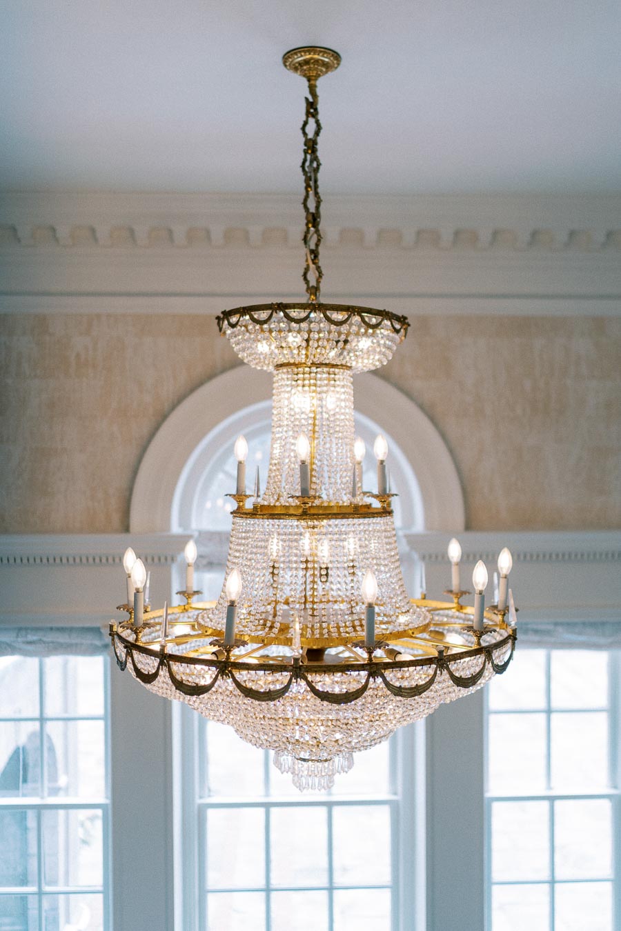 Elegant crystal chandelier with gold accents hanging from a ceiling, illuminated by multiple candle-shaped bulbs, with a background of large windows and arched details.