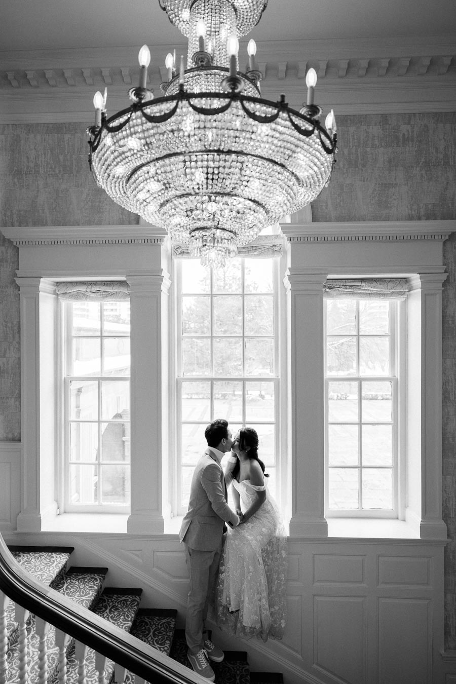 Black and white photo of a couple embracing under an elegant chandelier, with the woman sitting on a windowsill and the man standing on a staircase, creating a romantic atmosphere in a historic setting.