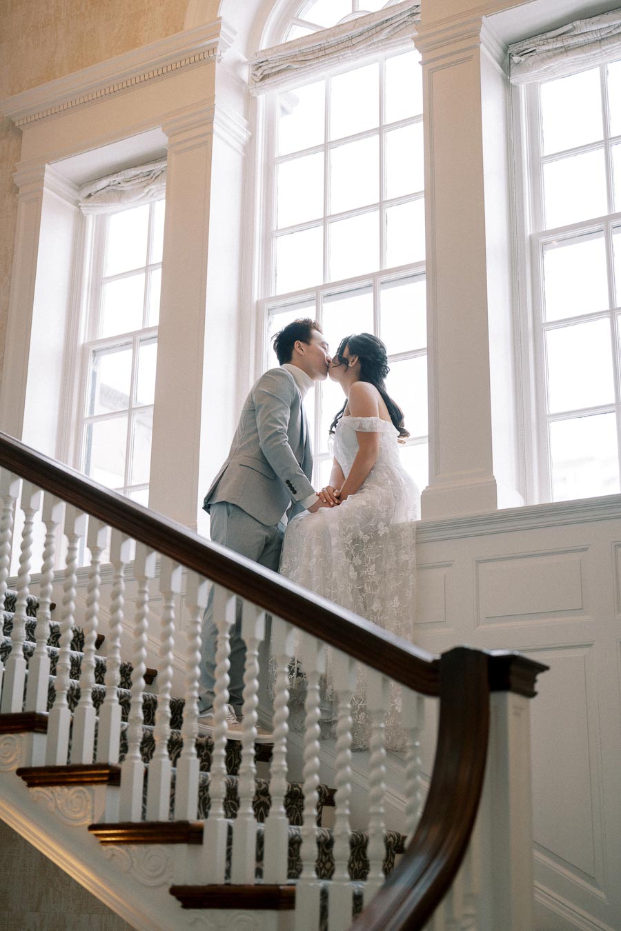 A couple shares a romantic kiss on a staircase by a large window, with elegant white railings and wall paneling, capturing a beautiful wedding moment in a classic, sophisticated setting.