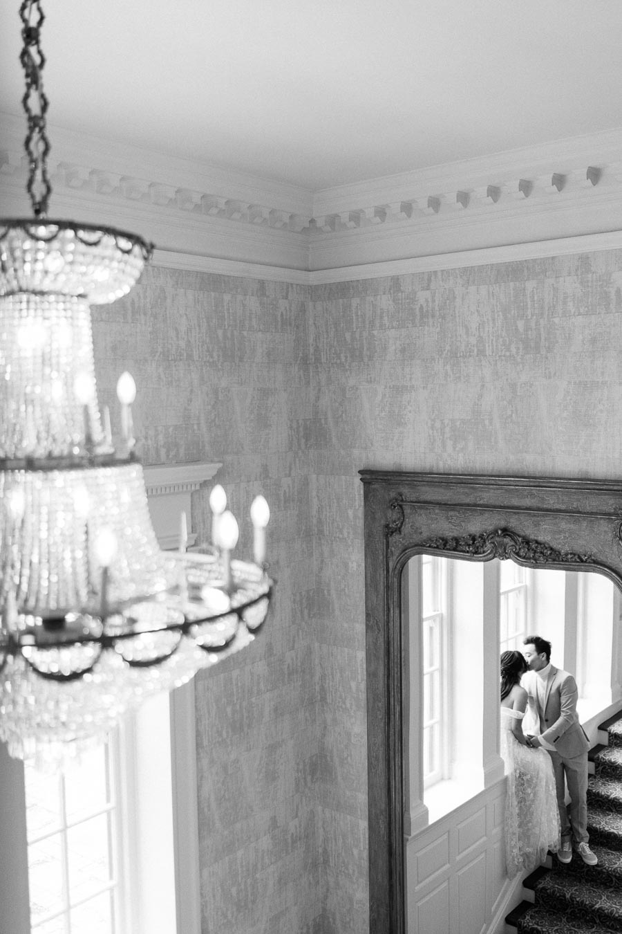Black and white photo of a couple embracing by a window inside an elegant room with a chandelier and ornate mirror, creating a romantic and timeless atmosphere.