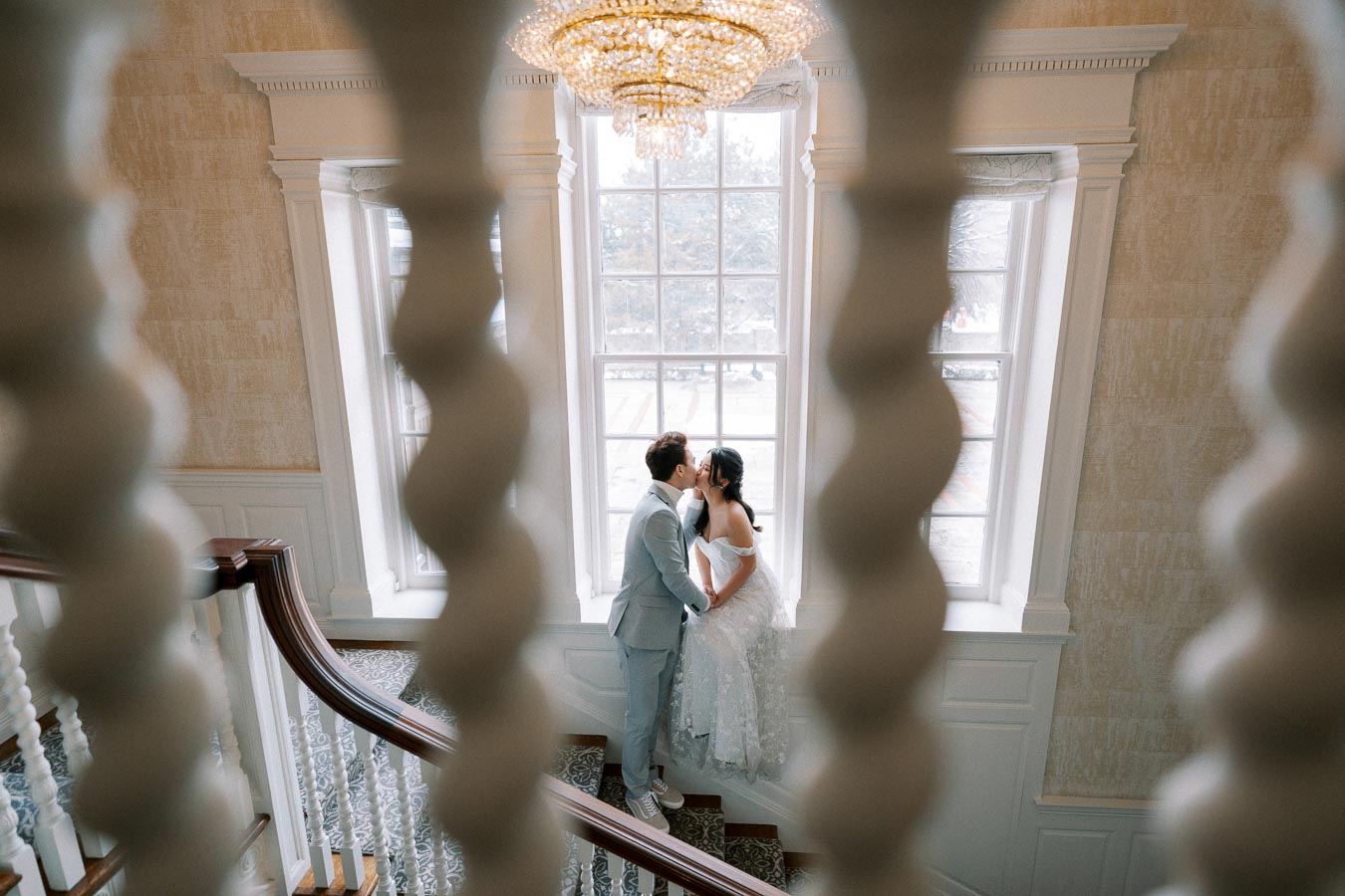 A couple in elegant attire share a romantic moment on a staircase framed by ornate railing, with a chandelier above and a large window in the background.