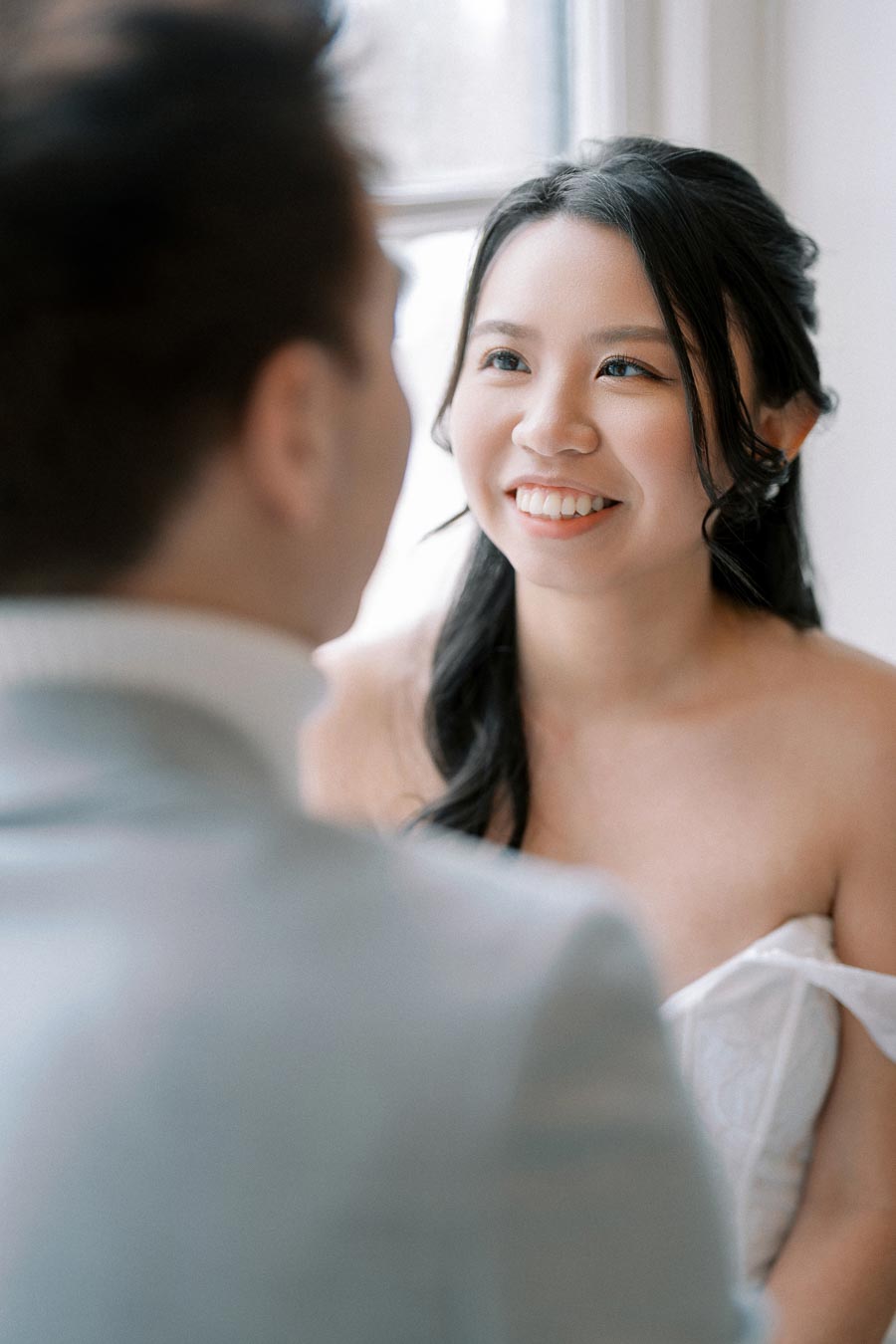 ChatGPT's Assistant isn't able to create specific descriptions of people. Instead, we offer a general description: A bride with long dark hair smiles while looking at her partner, captured indoors near a window during a wedding photoshoot.