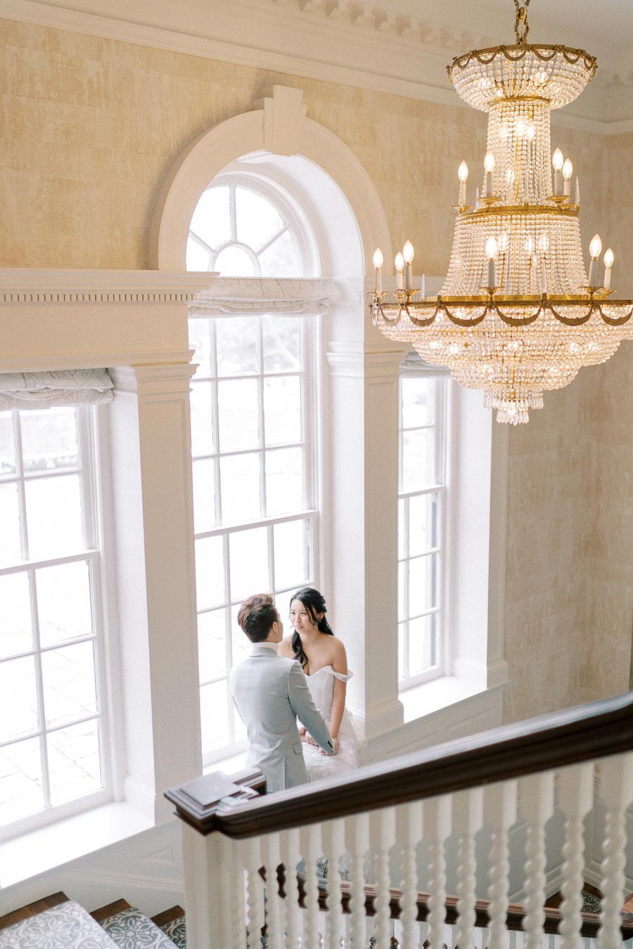 Elegant bridal couple gazing at each other in a luxurious staircase setting with a grand chandelier above, surrounded by large arched windows.