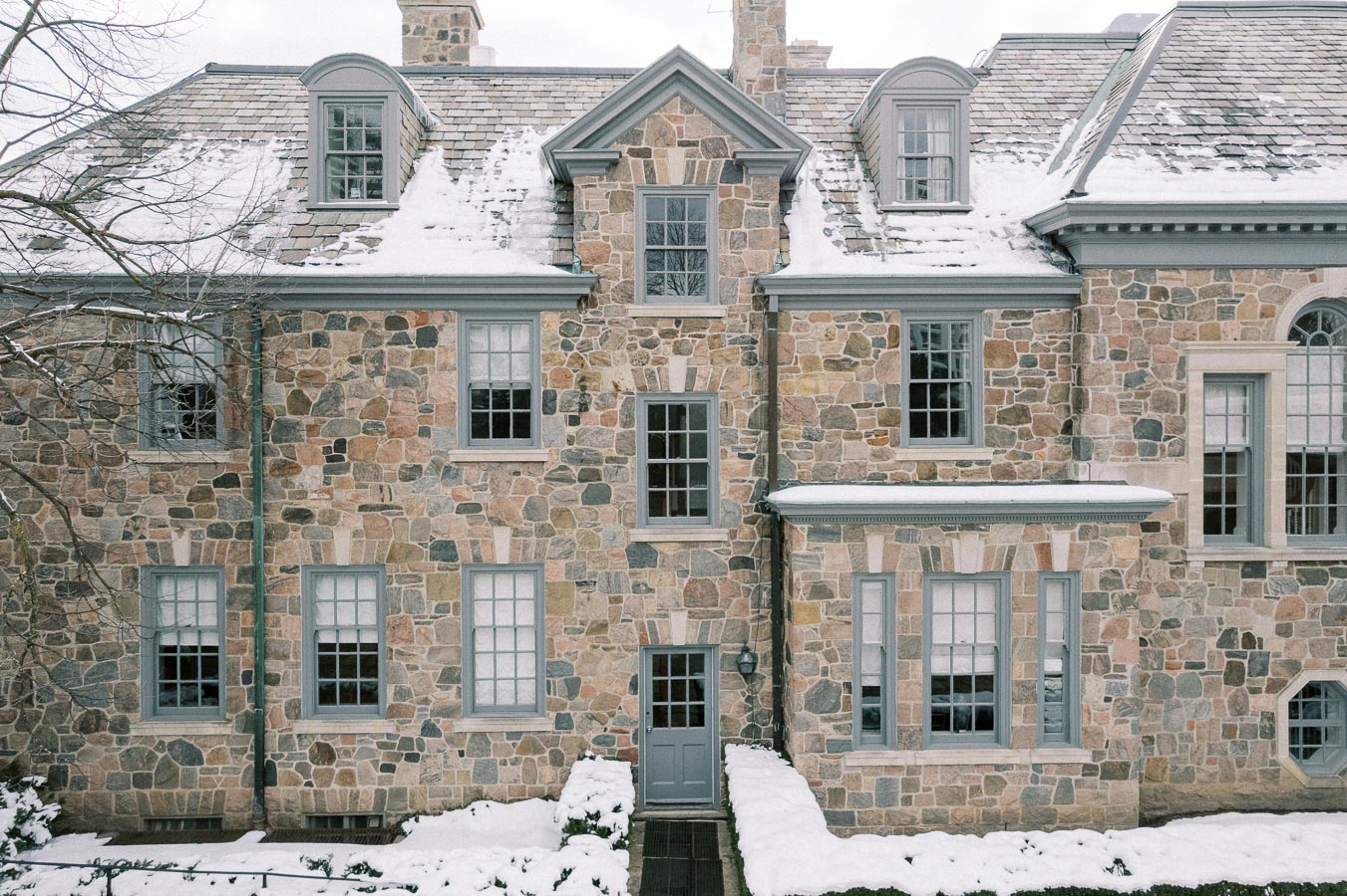 Stone facade of a traditional house with snow-covered roof and window sills, showcasing classic architecture in a winter landscape.