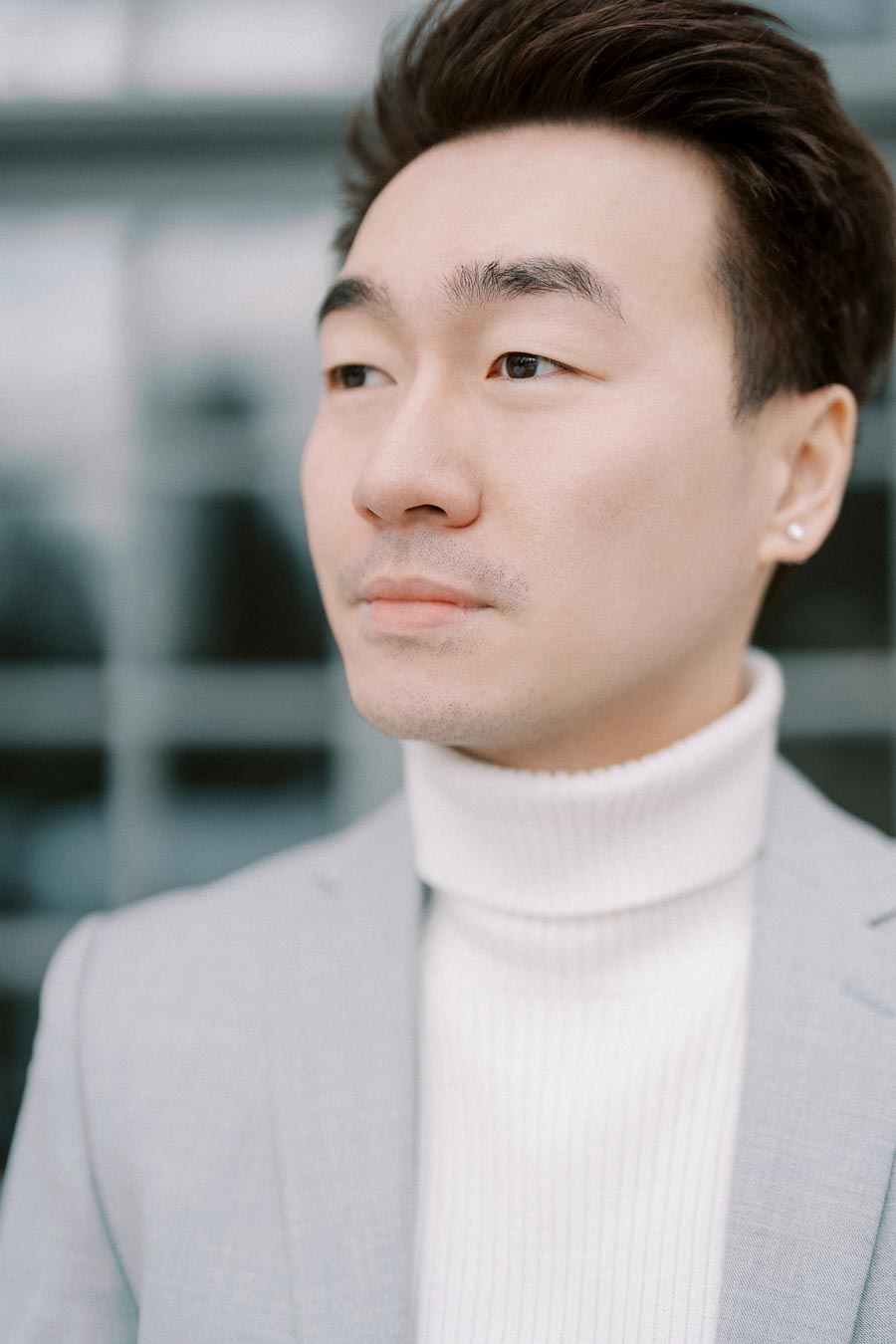 A stylish individual gazes thoughtfully into the distance, wearing a light gray blazer and white turtleneck, set against a blurred background.