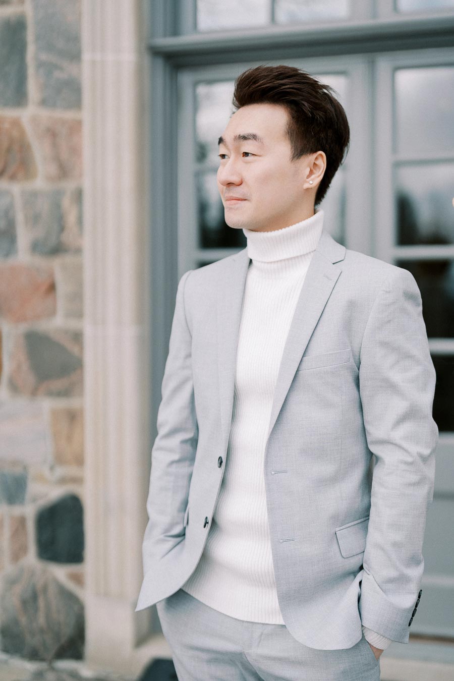 A man in a light gray suit and white turtleneck stands confidently in front of a stone building with large windows, showcasing modern fashion and style.
