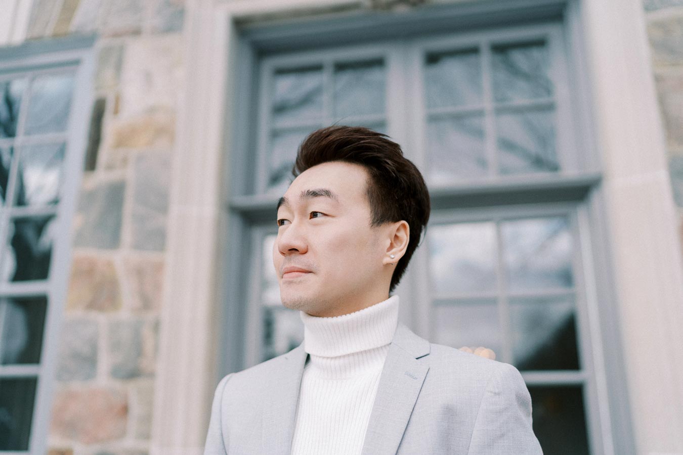A person in a light gray suit and white turtleneck stands outside, looking thoughtful against a backdrop of a stone building with large windows.