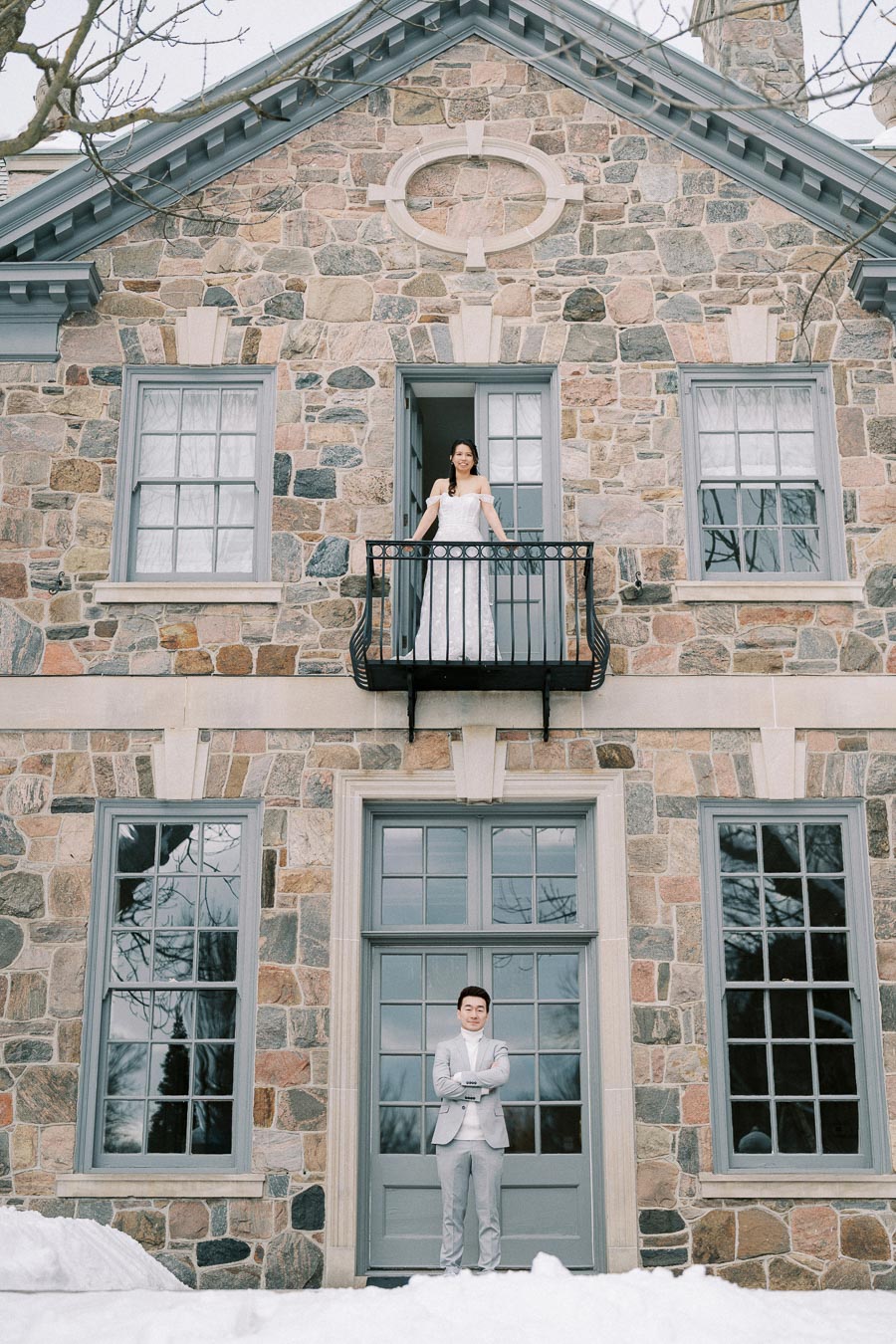 Bride and groom standing outside a rustic stone building, with the bride on a small balcony above and the groom below on the snowy ground, showcasing a winter wedding scene.
