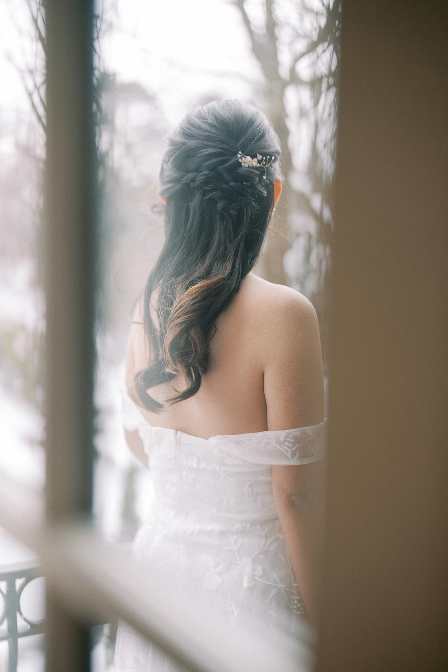 Bridal portrait from behind, showcasing an elegant off-shoulder lace wedding dress and beautifully styled dark hair adorned with a decorative clip, set against a blurred background of trees.