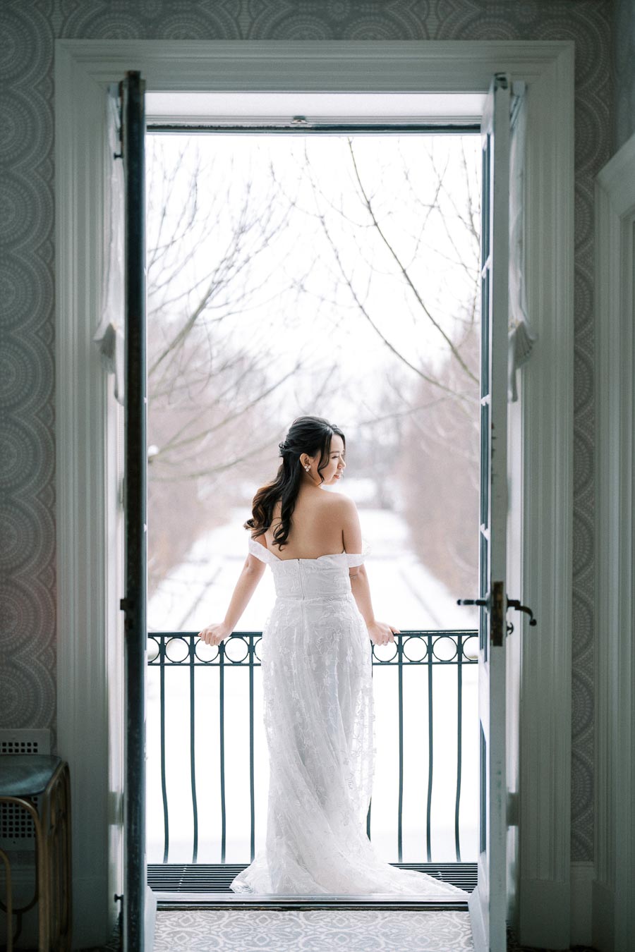 Certainly! Here's an SEO-optimized alt text:Elegant bride in white lace dress standing on a balcony overlooking a snowy winter landscape, framed by an open doorway.