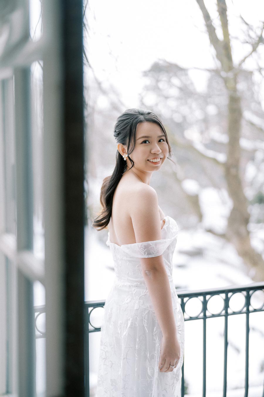A bride in a white lace wedding dress smiles while standing on a balcony during winter, with snow-covered trees in the background.