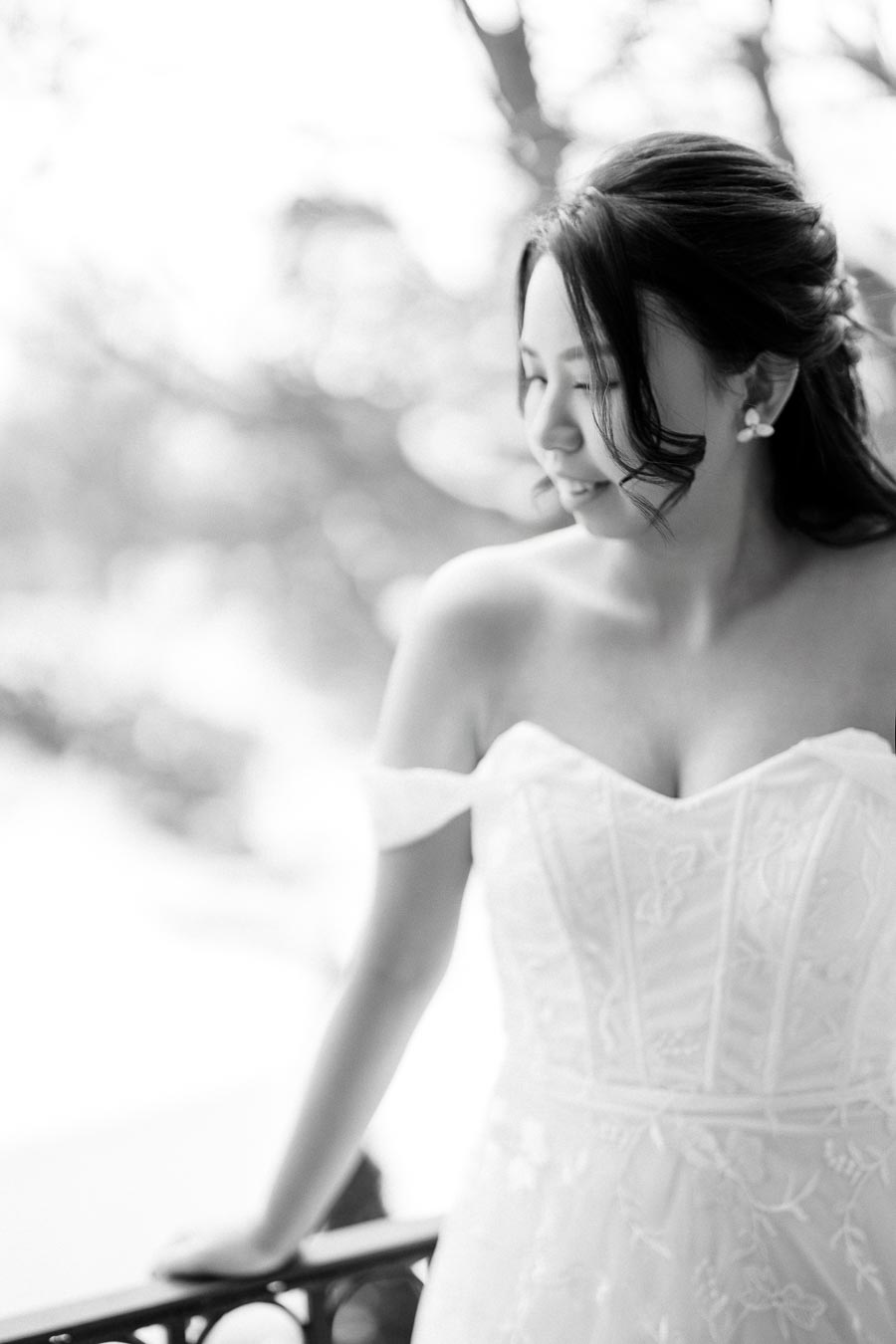 A bride in an elegant white wedding dress standing on a balcony, captured in soft focus black and white photography, showcasing a serene and joyful expression.