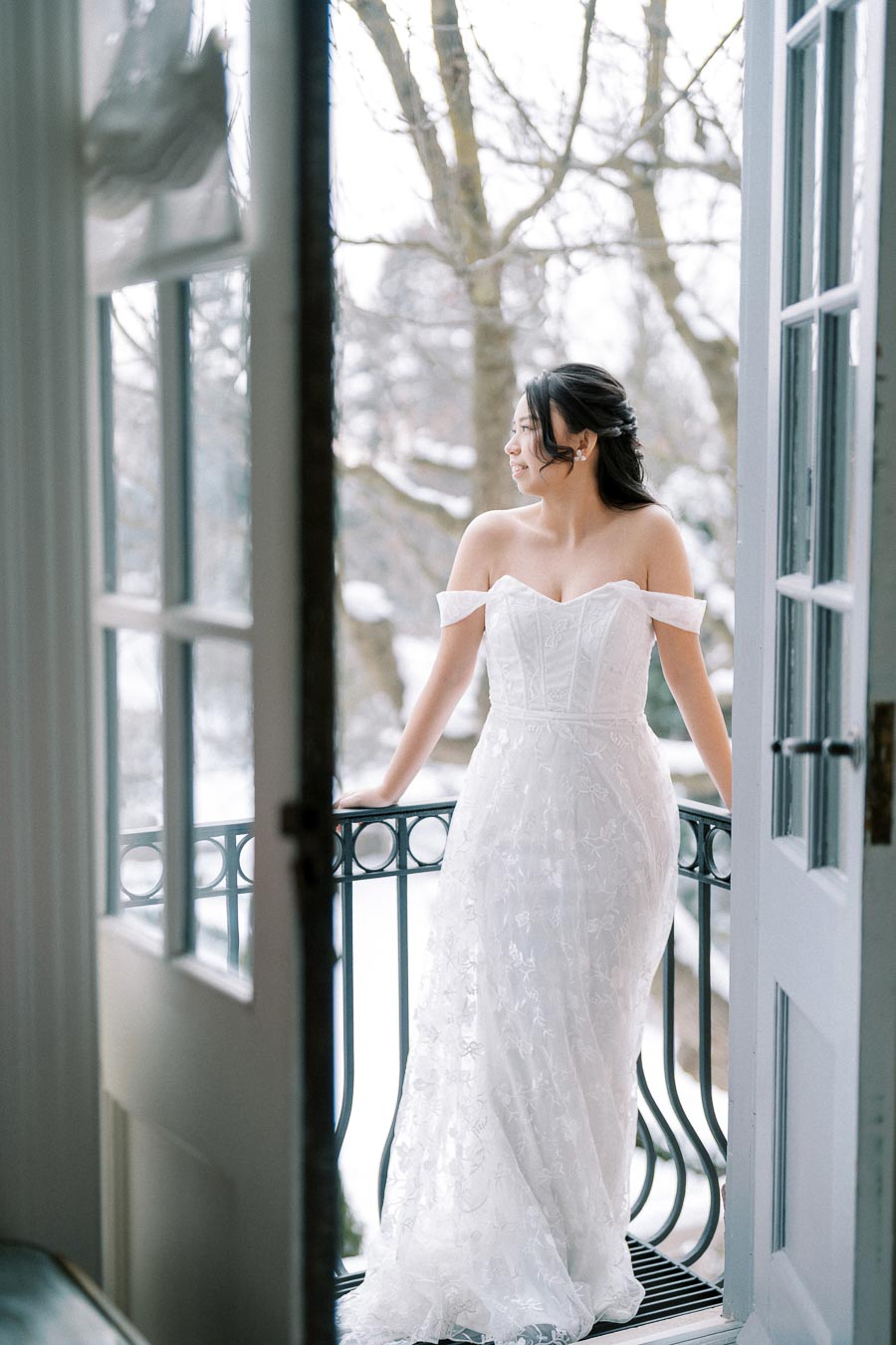 Bride in elegant off-shoulder wedding dress standing on a balcony, with a serene snowy landscape in the background.