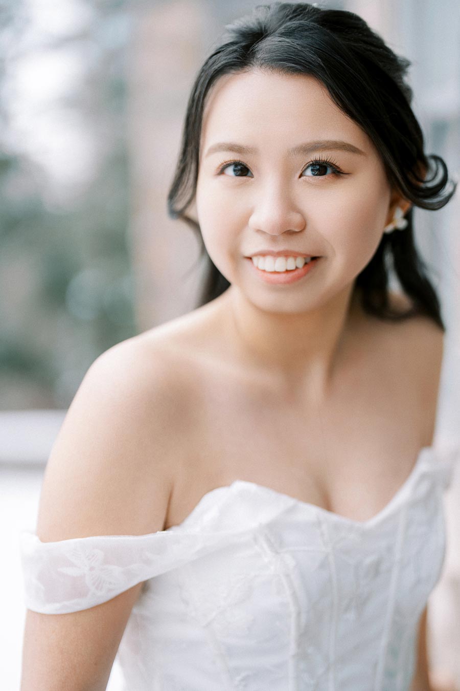 Alt text: Smiling bride in an elegant, off-the-shoulder white wedding dress, looking directly at the camera with soft, natural lighting.