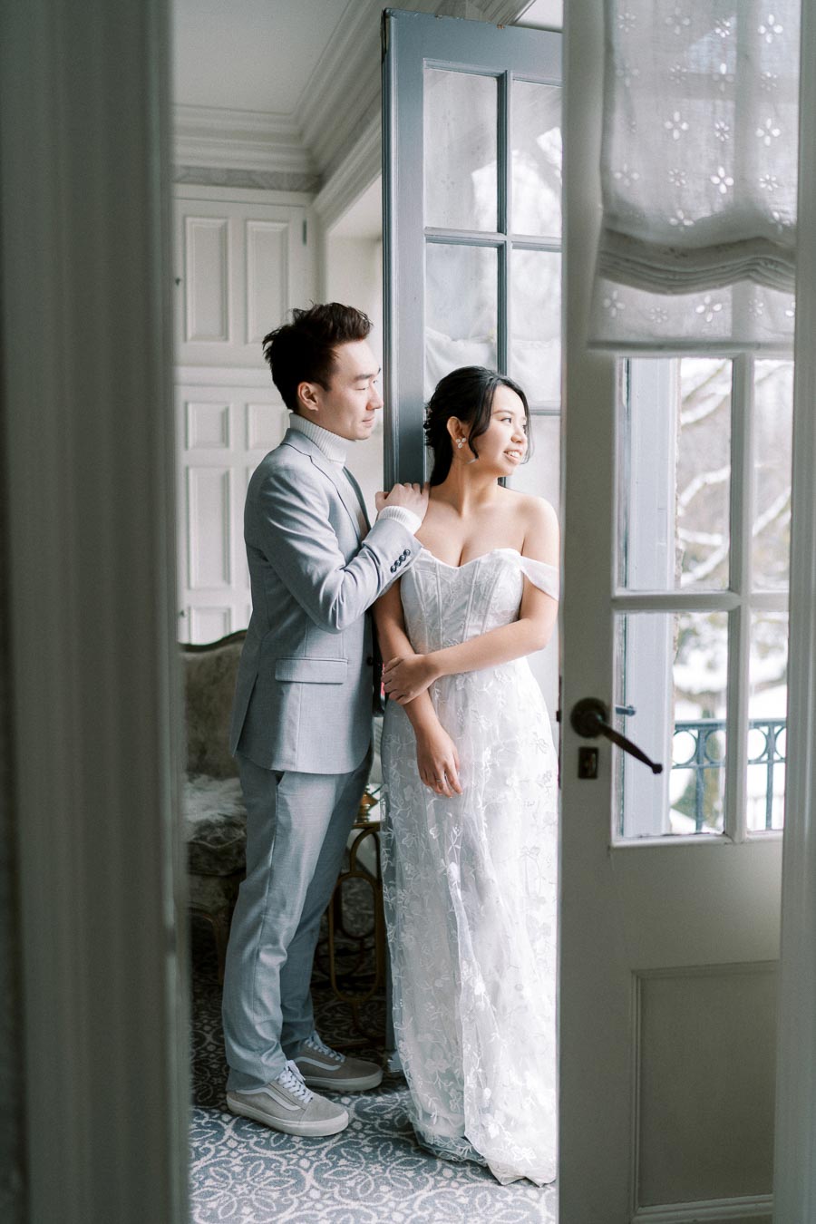A couple stands elegantly by a French door, gazing outside on a snowy day; the woman is in a white bridal gown while the man wears a light gray suit, creating a serene and romantic ambiance in a softly lit room.