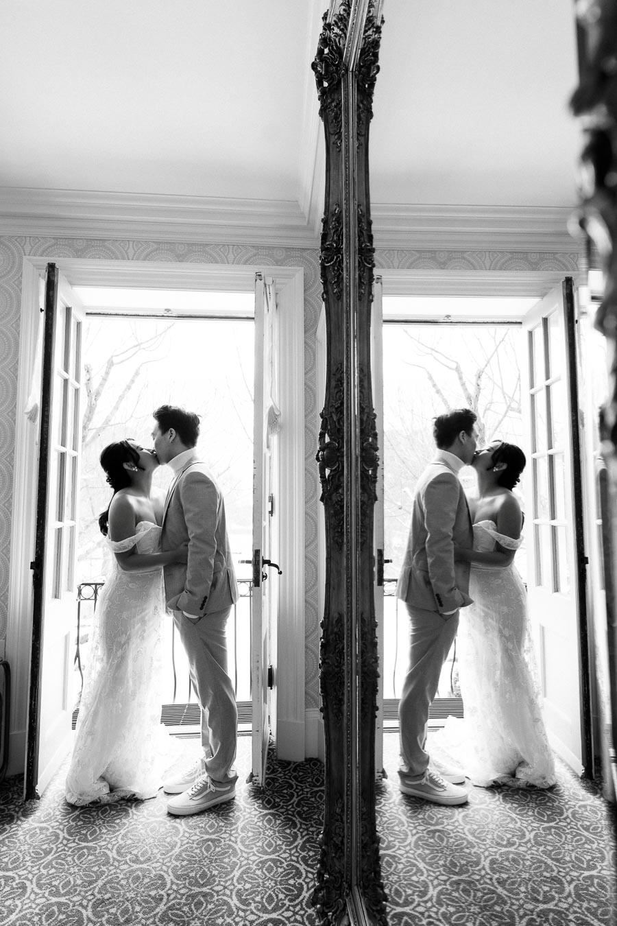 Black and white photo of a couple sharing a kiss by a large window, with their reflection visible in a decorative mirror, capturing a romantic indoor wedding moment.