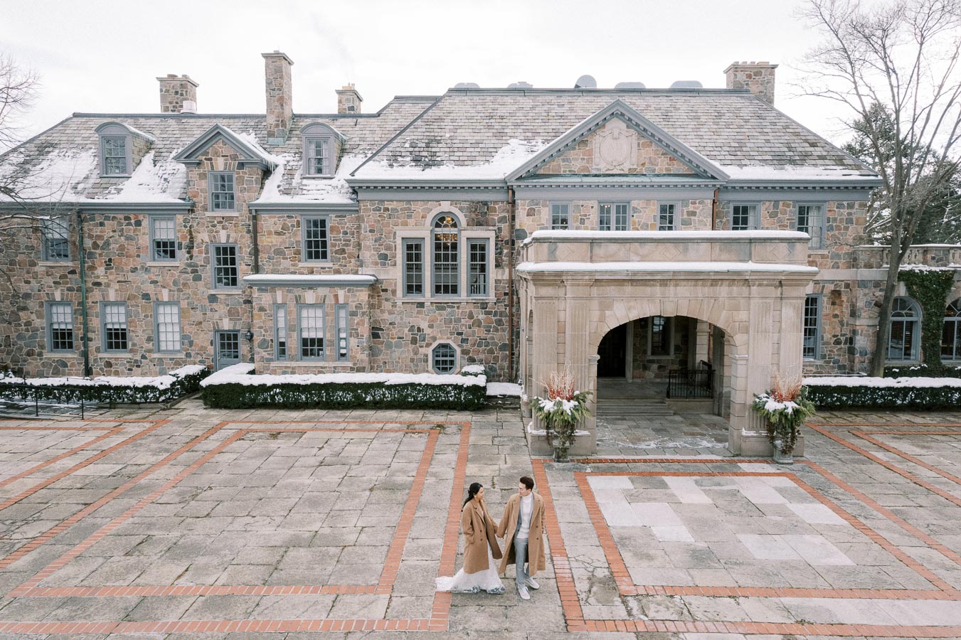 Elegant couple holding hands in front of a historic stone mansion with snow on the roof, showcasing winter romance and architectural beauty.