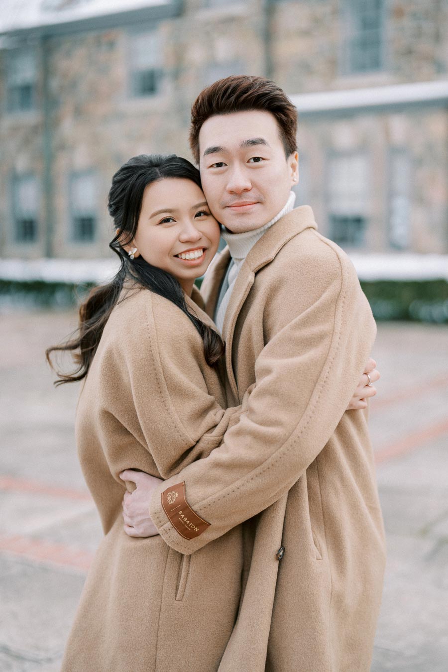 A couple embracing outdoors in winter coats, standing in front of a historic stone building.