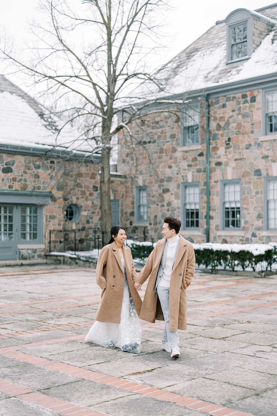 A couple in matching beige coats holding hands and smiling while walking on a snowy stone pathway in front of a historic stone building.
