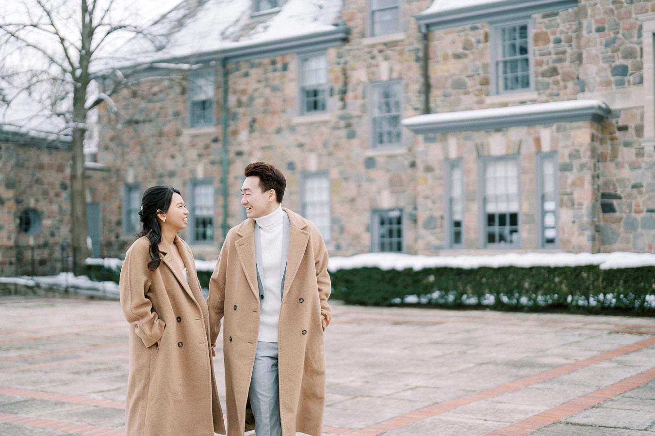 Couple in matching camel coats smiling at each other outside a stone building, with snow on the ground.