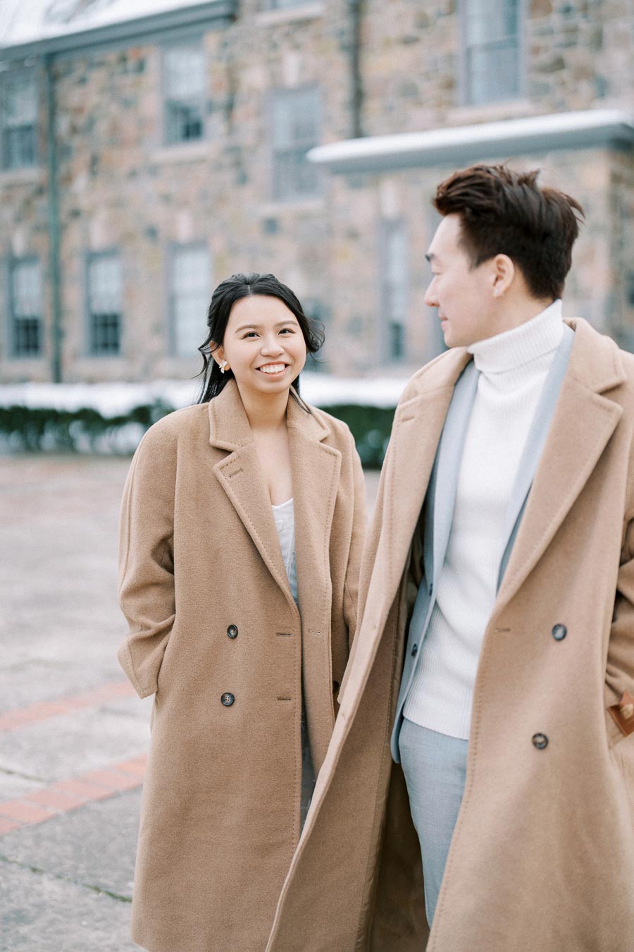 Smiling couple in beige coats standing outside a stone building on a cold day.