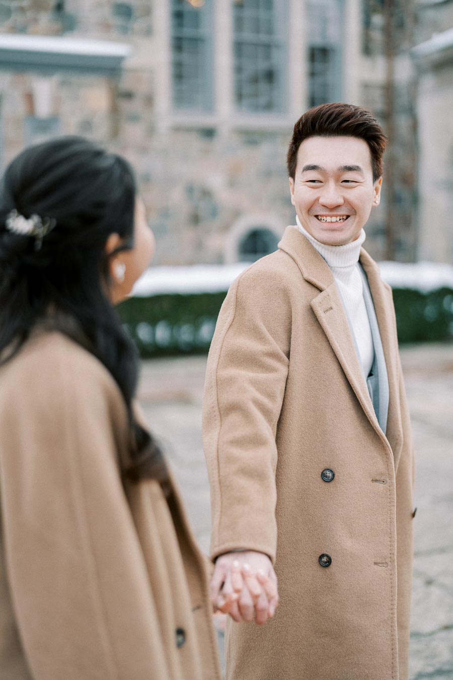 A couple holding hands and smiling, dressed in matching beige coats, standing in front of a stone building.