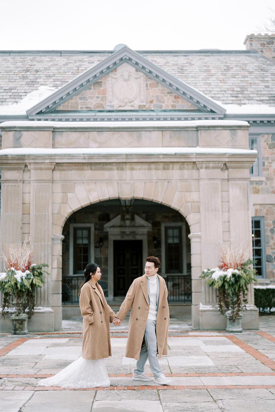 Couple holding hands in stylish winter coats in front of a historic stone building, showcasing elegant winter fashion and romantic architecture.