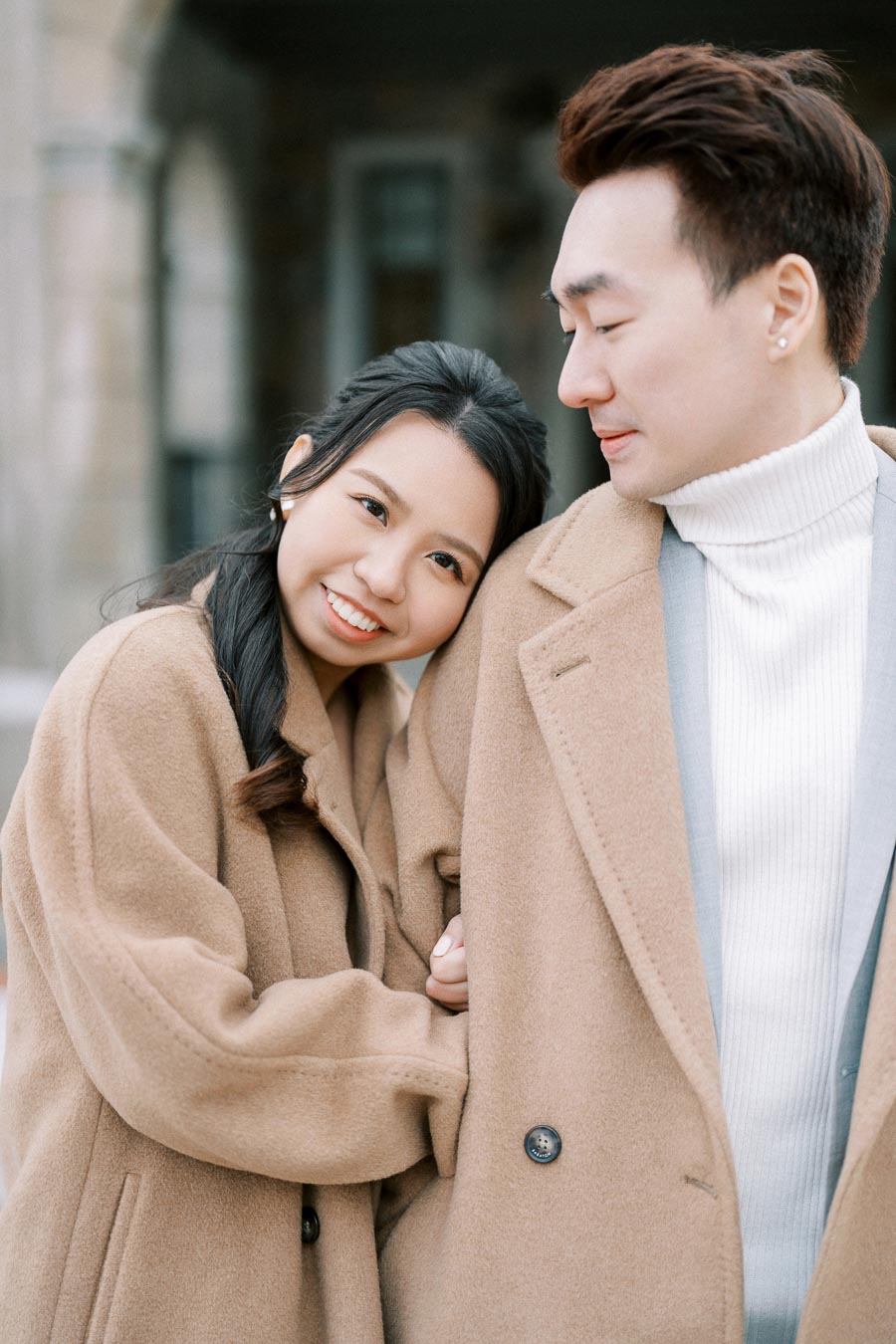 A happy couple in beige winter coats displaying affection outdoors, with the woman smiling warmly and leaning on the man's arm.
