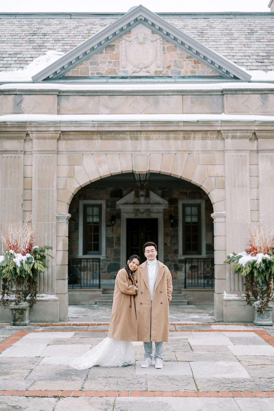 A couple in matching beige coats poses in front of a historic stone building during winter, with snow visible on the ground and roof, creating a romantic and elegant atmosphere.