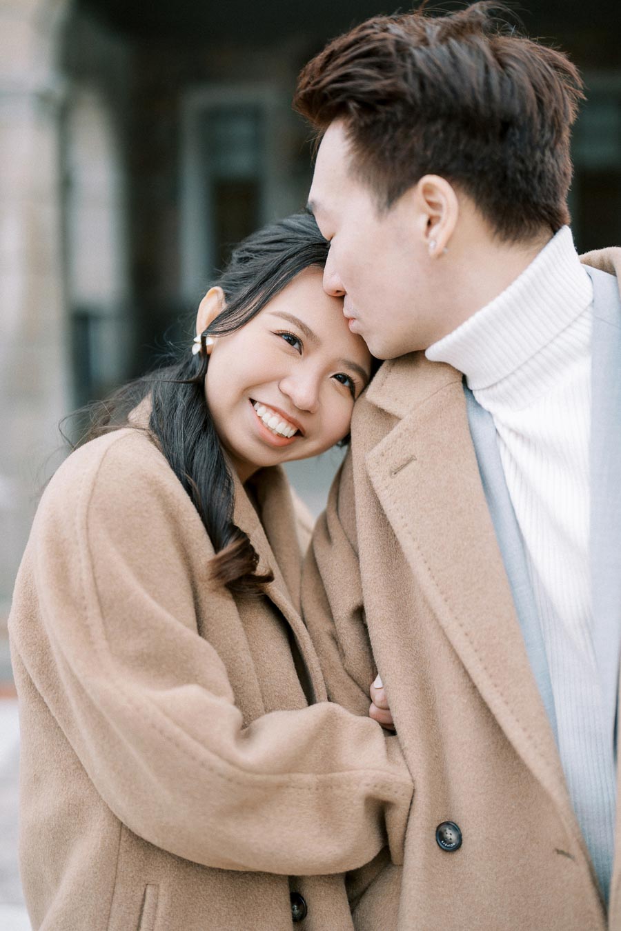 A couple in matching brown coats sharing an intimate moment, with the man gently kissing the woman's forehead as she smiles warmly, showcasing a loving and tender connection.