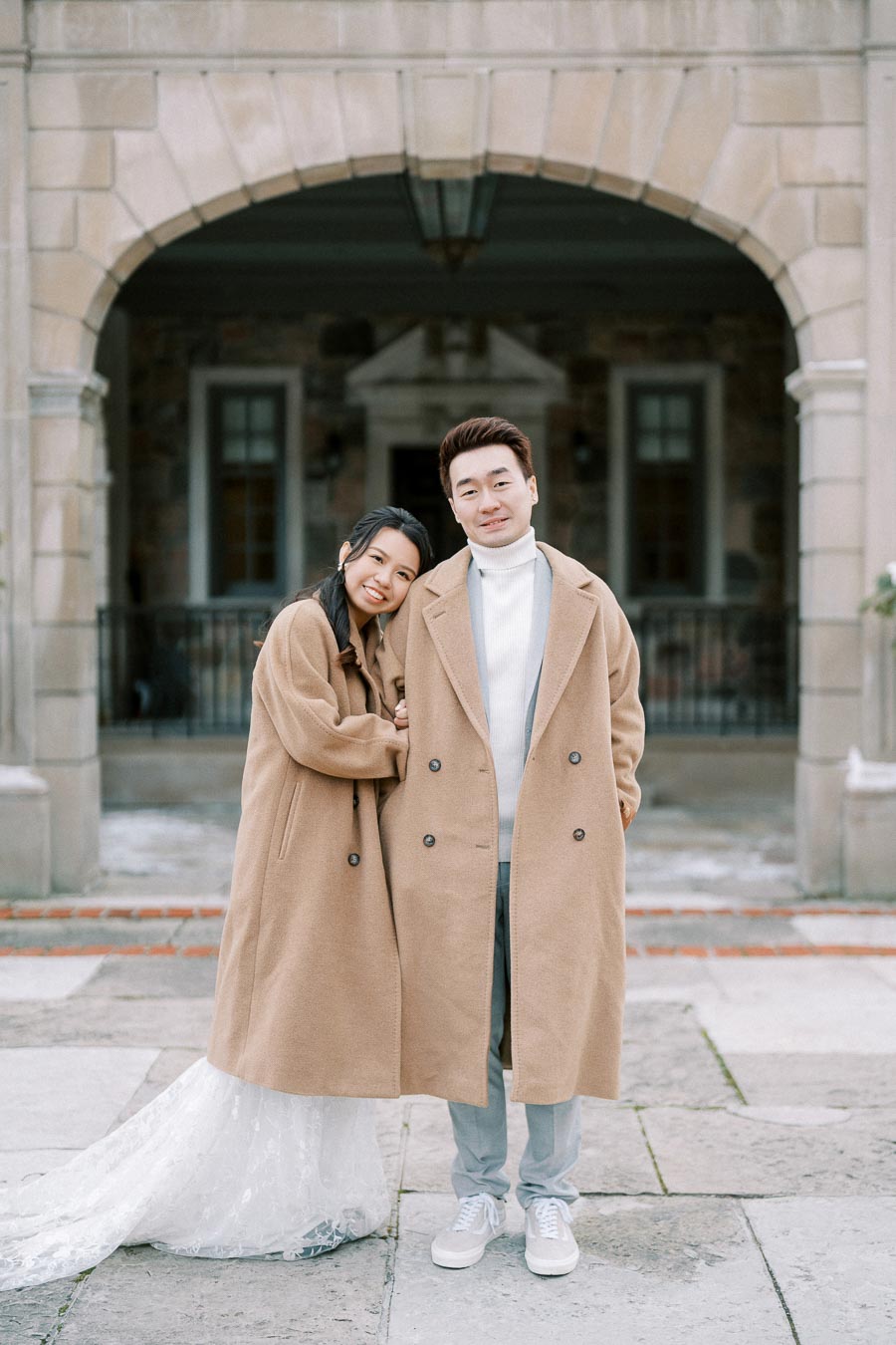 Couple posing in matching beige coats, standing in front of an elegant stone building. The woman rests her head on the man's shoulder, wearing a white lace dress underneath her coat. The man wears a white turtleneck and light pants, both smiling softly.