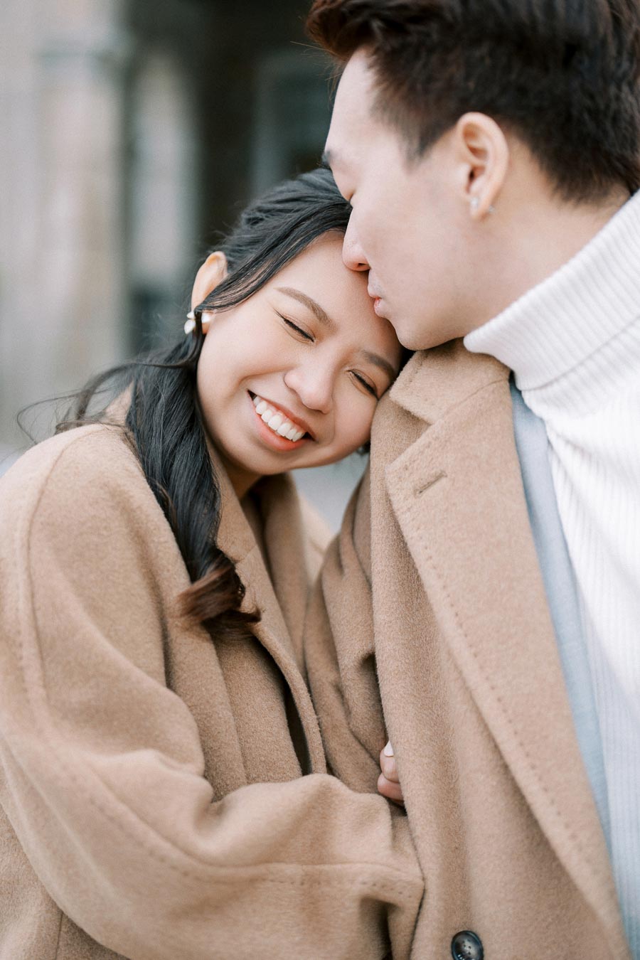A couple in matching beige coats shares a tender moment, with the woman smiling and resting her head on the man's shoulder outdoors.