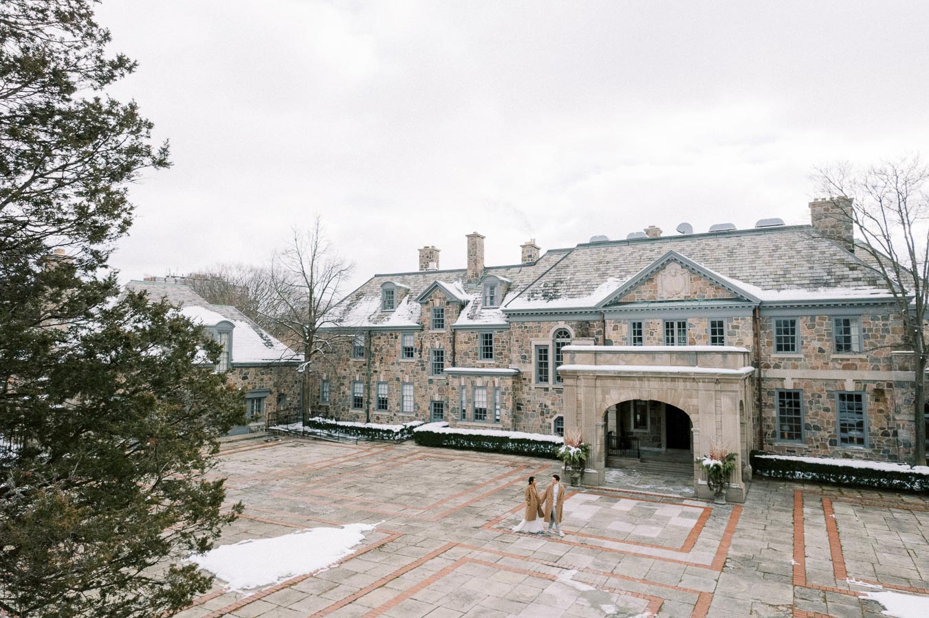 Elegant stone manor house with a snow-dusted roof, featuring large windows and an arched entrance. A couple dressed in winter attire walk across the spacious cobblestone courtyard, surrounded by bare trees on a cloudy day.