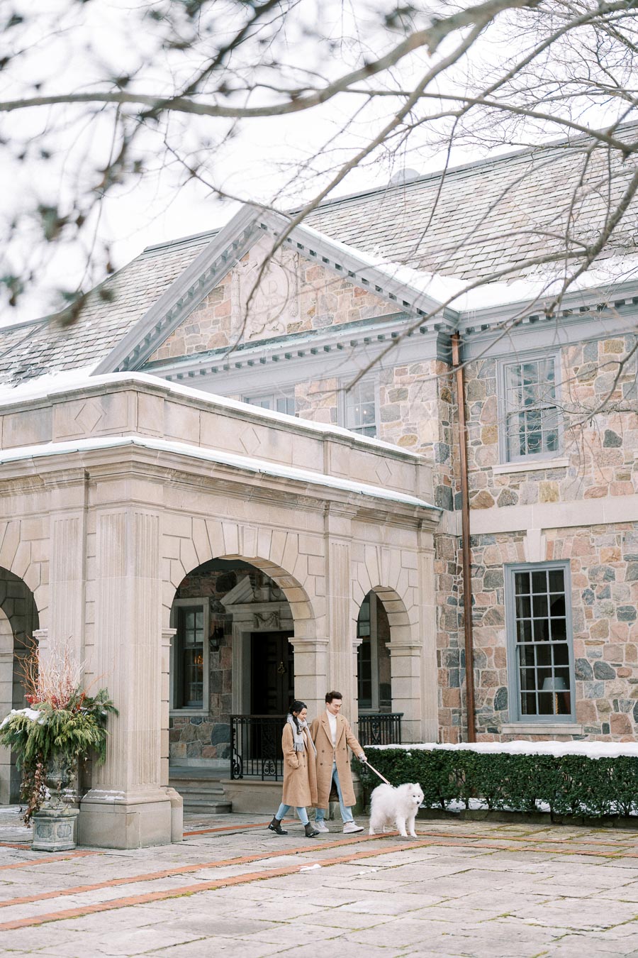 Elegant stone mansion with arched entry, a couple in matching coats walks their fluffy white dog on the snowy pathway in early winter.
