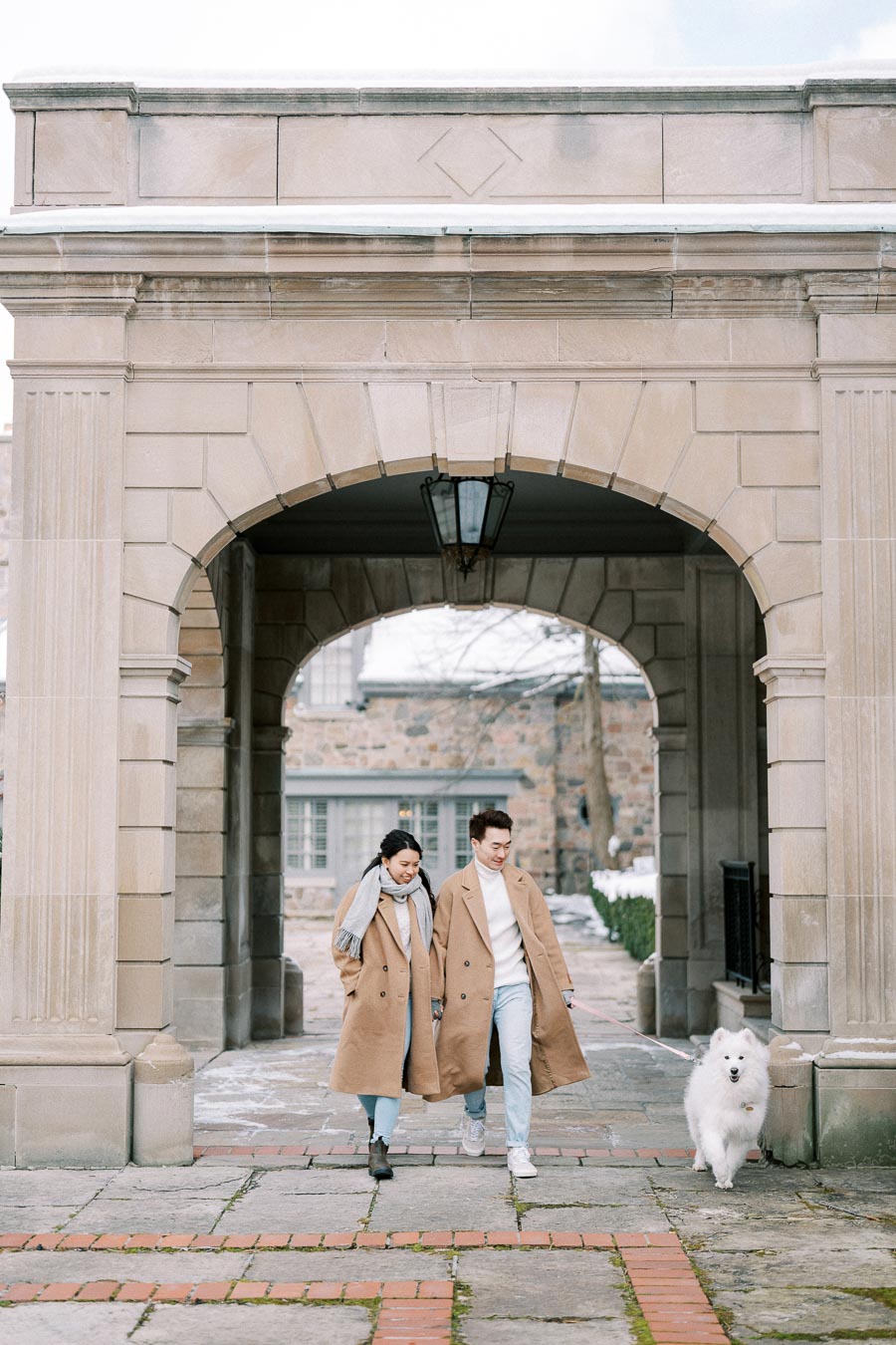 A couple in matching beige coats walks a fluffy white dog through a stone archway on a winter day.