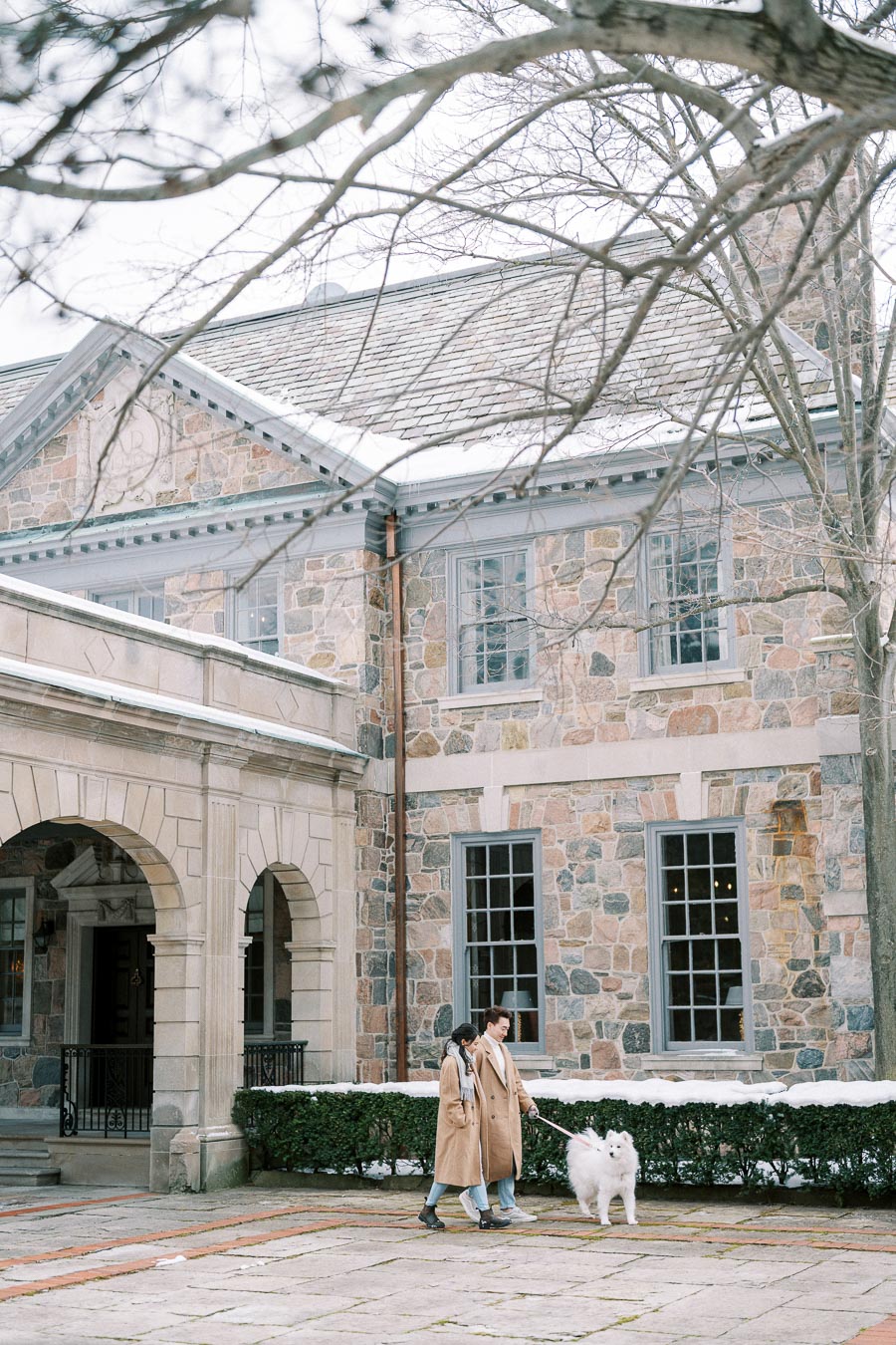 Couple walking their fluffy white dog in front of a stone building with arched entrance and snow-dusted surroundings.