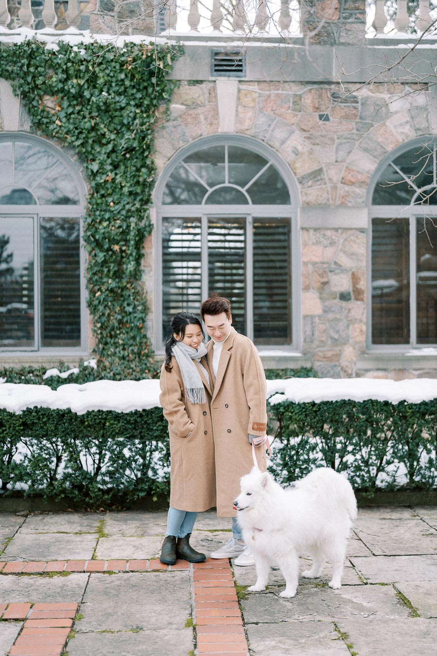 A couple in matching beige coats stands closely together on a snowy patio, smiling affectionately at each other. They are accompanied by a fluffy white dog, with ivy growing on a stone building in the background.