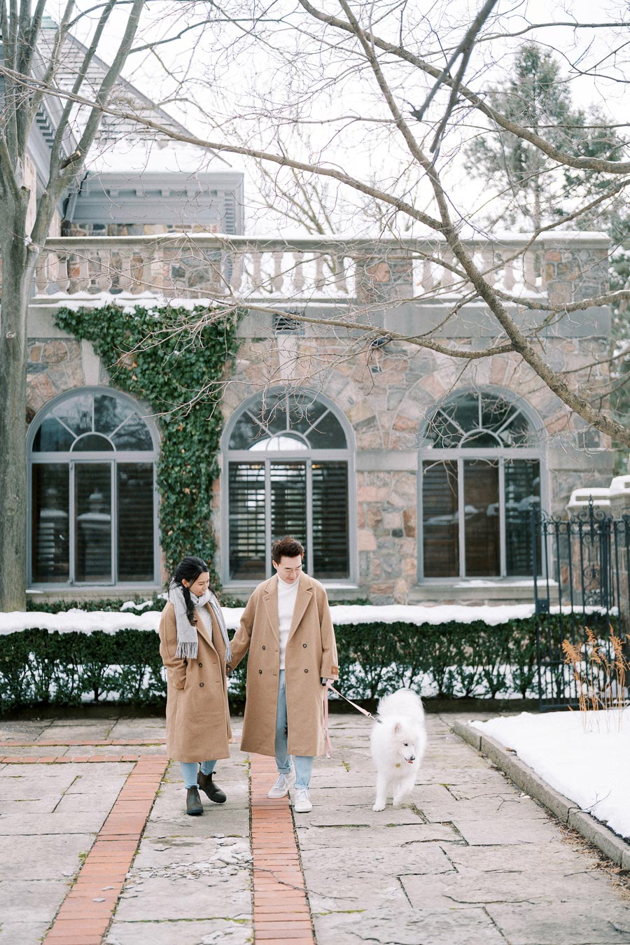 A couple in matching tan coats walks their fluffy white dog in a snowy courtyard in front of a stone building with ivy-covered walls and arched windows.