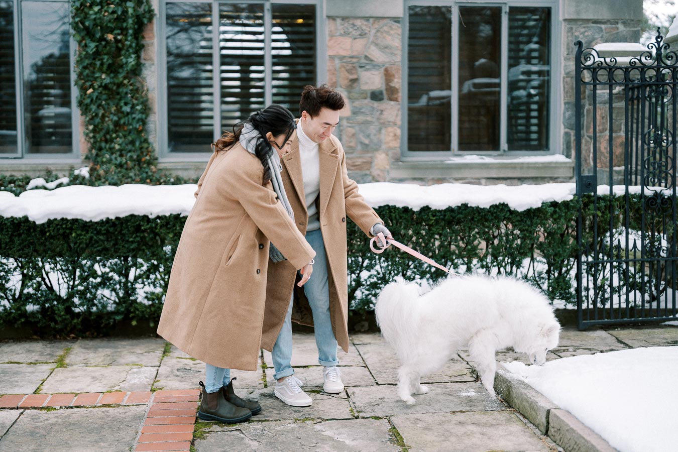 Couple in matching tan coats walking their fluffy white dog on a snowy sidewalk, with a brick house and greenery in the background.