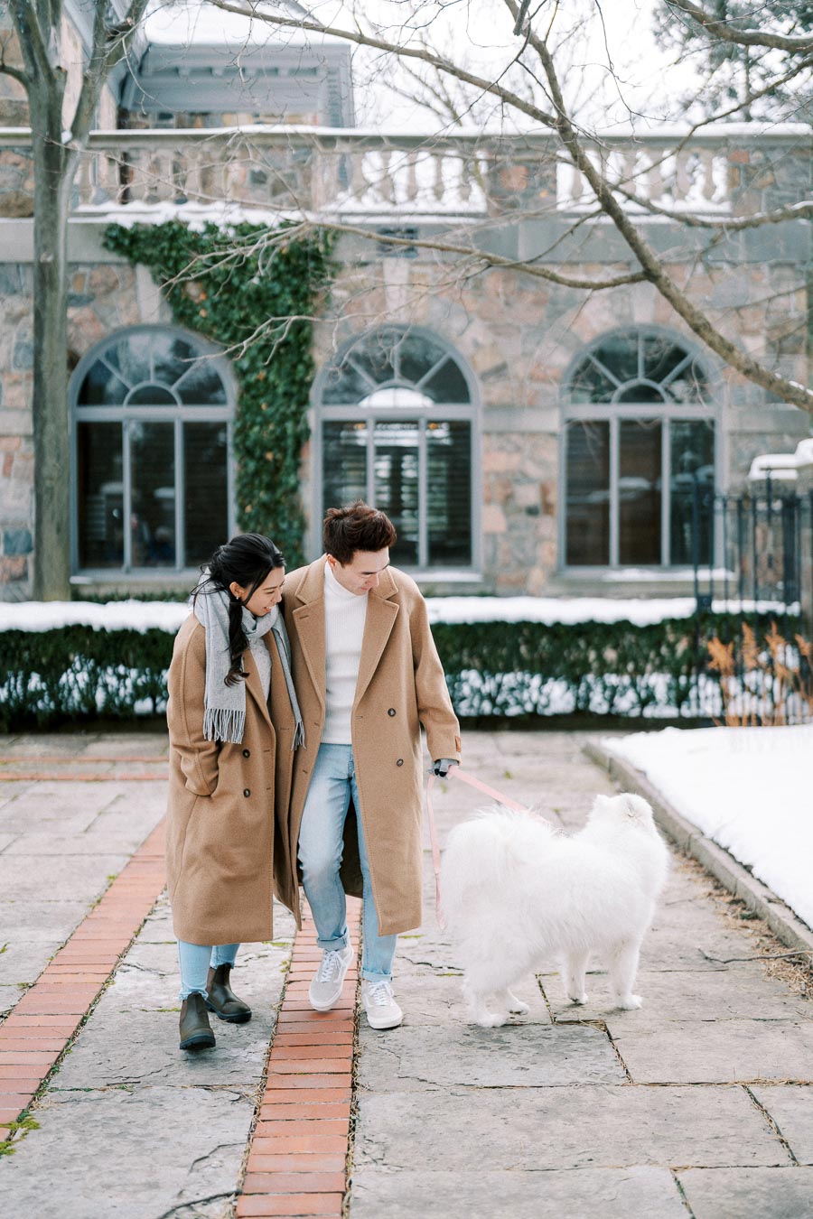 A couple in matching beige coats strolling with a white fluffy dog on a snowy path in front of a stone building with arched windows, creating a cozy winter scene.