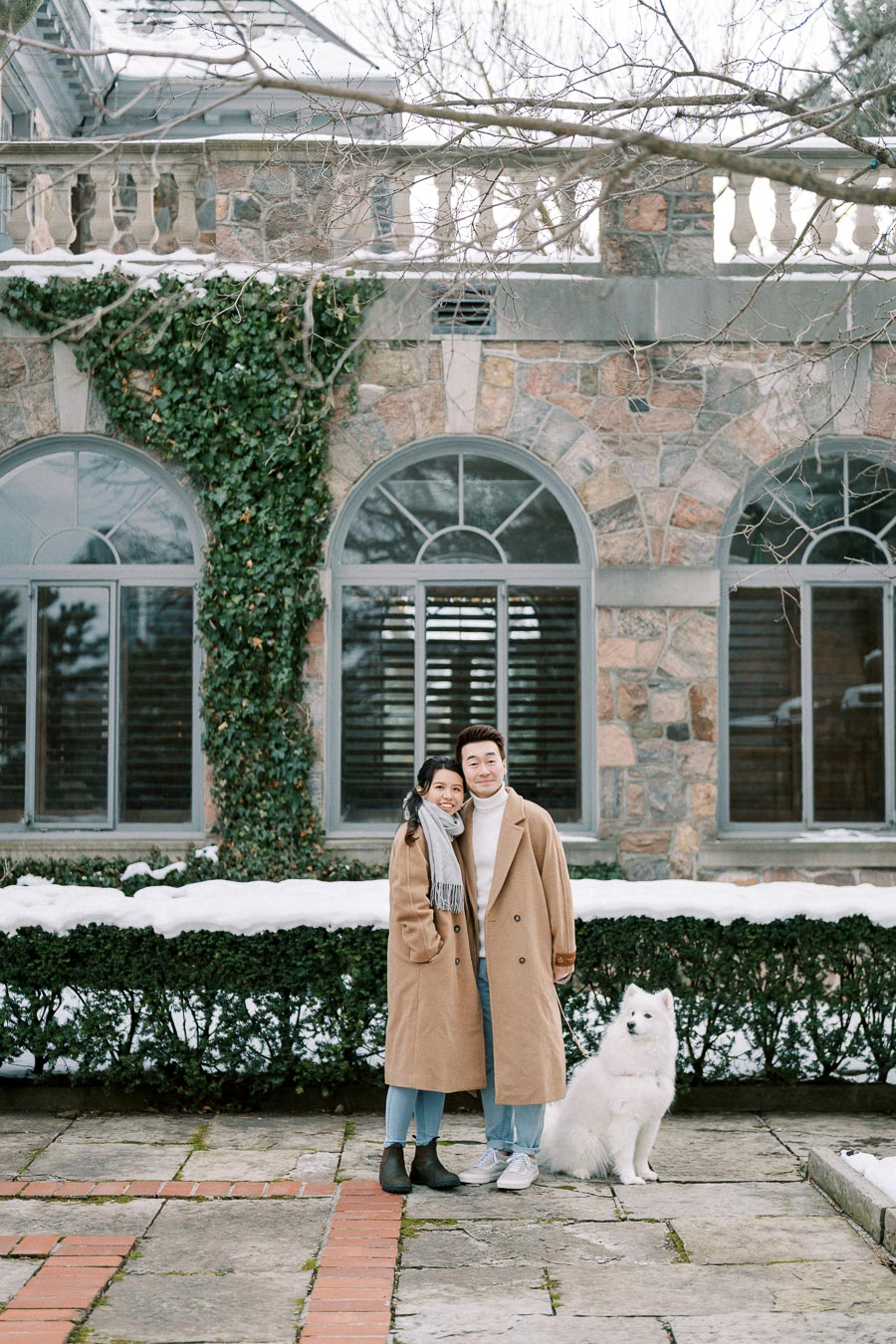A couple in matching beige coats smiling in front of a historic stone building with arched windows, accompanied by a fluffy white dog, with snow on the ground and ivy climbing the walls.