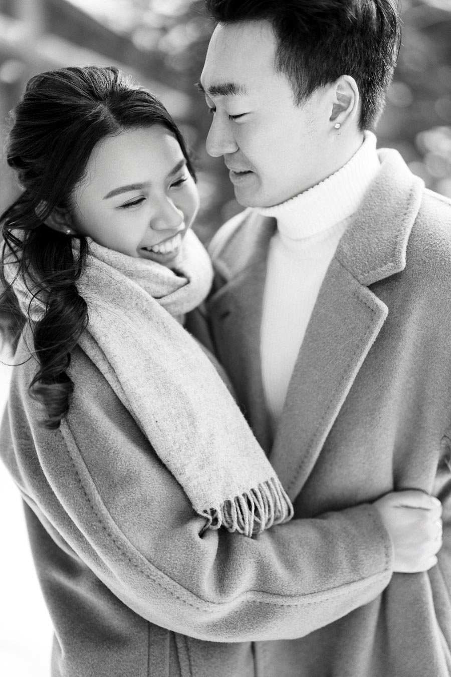 A black and white photo of a smiling couple embracing warmly, both wearing winter coats and scarves, exuding happiness and love in a cozy outdoor setting.