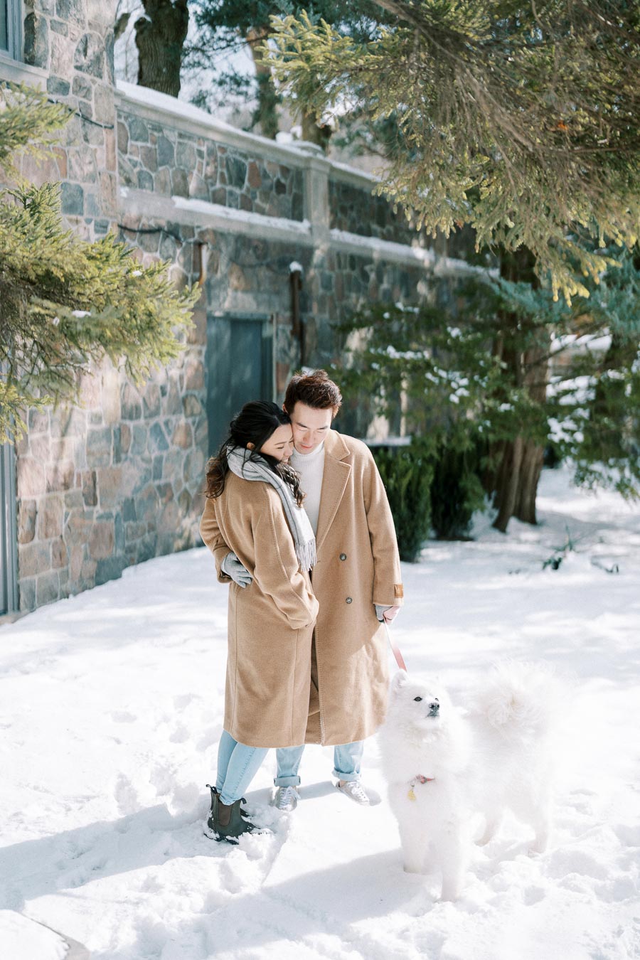 A couple dressed in matching tan coats embraces in a snowy landscape with a fluffy white dog on a leash, near a stone building and surrounded by evergreen trees.