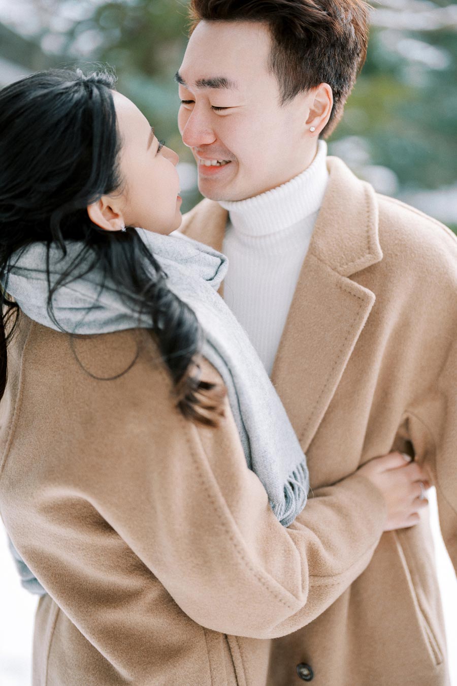 A couple wearing beige coats smiles at each other in a winter setting with green trees in the background.