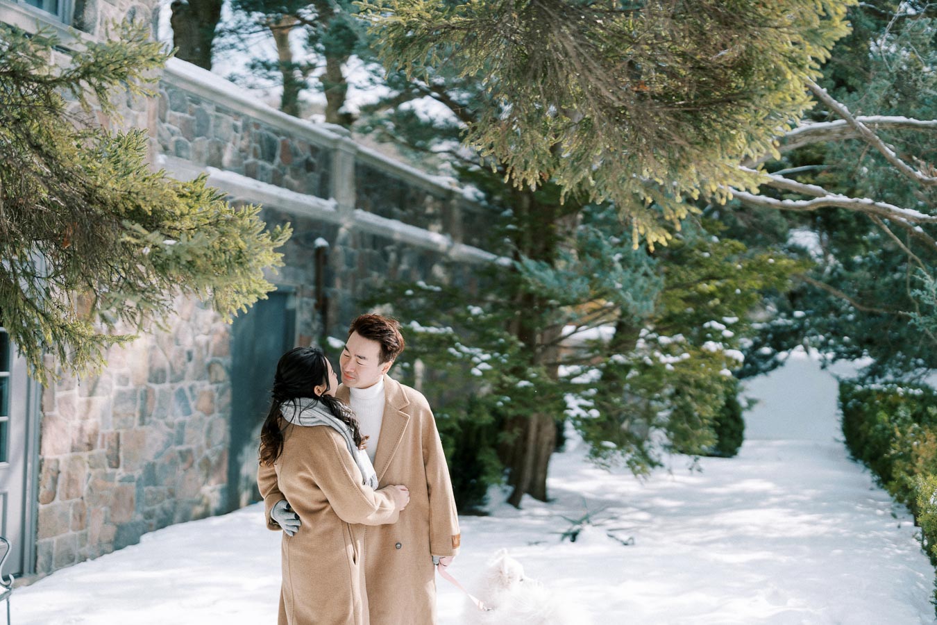 A couple embracing next to a stone building in a snowy forest setting, both wearing camel-colored coats and a scarf, with a fluffy white dog on a leash by their side.