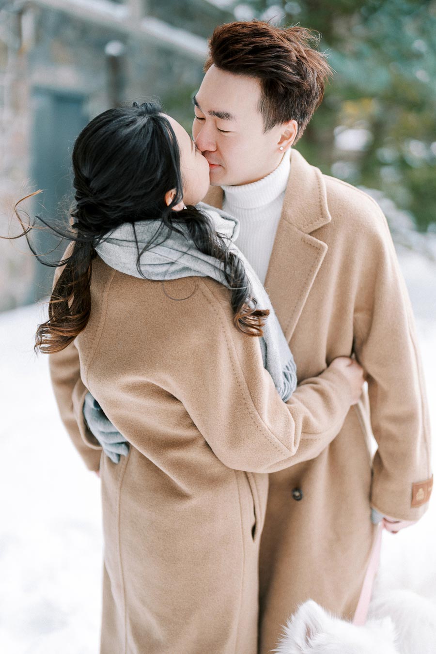 A couple embracing and kissing in a snowy winter landscape, both wearing tan coats and a scarf, with a fluffy white dog nearby.