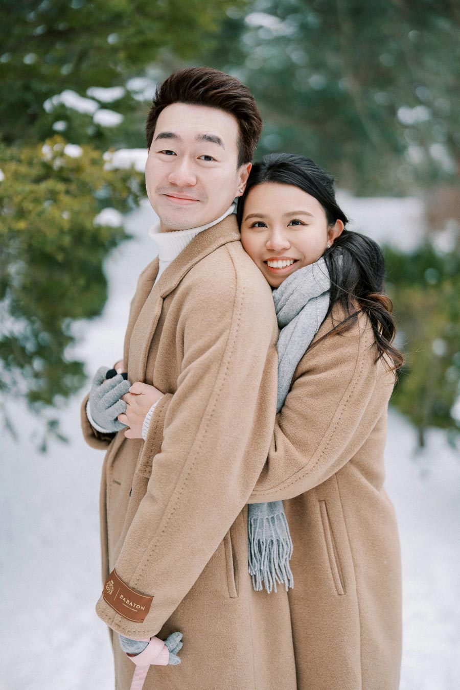 Couple embracing in snowy forest wearing matching camel coats, showcasing winter fashion and love in nature.
