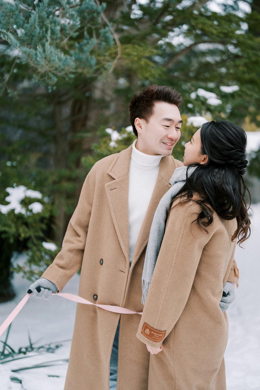 A couple in matching winter coats shares a joyful moment in a snowy forest, holding hands and smiling warmly at each other.