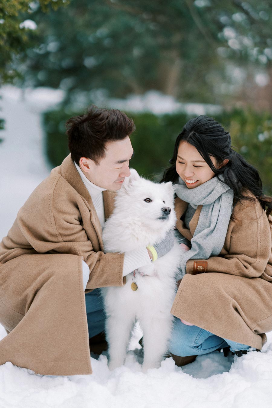 A couple in tan coats smiling and hugging a fluffy white dog in a snowy park.