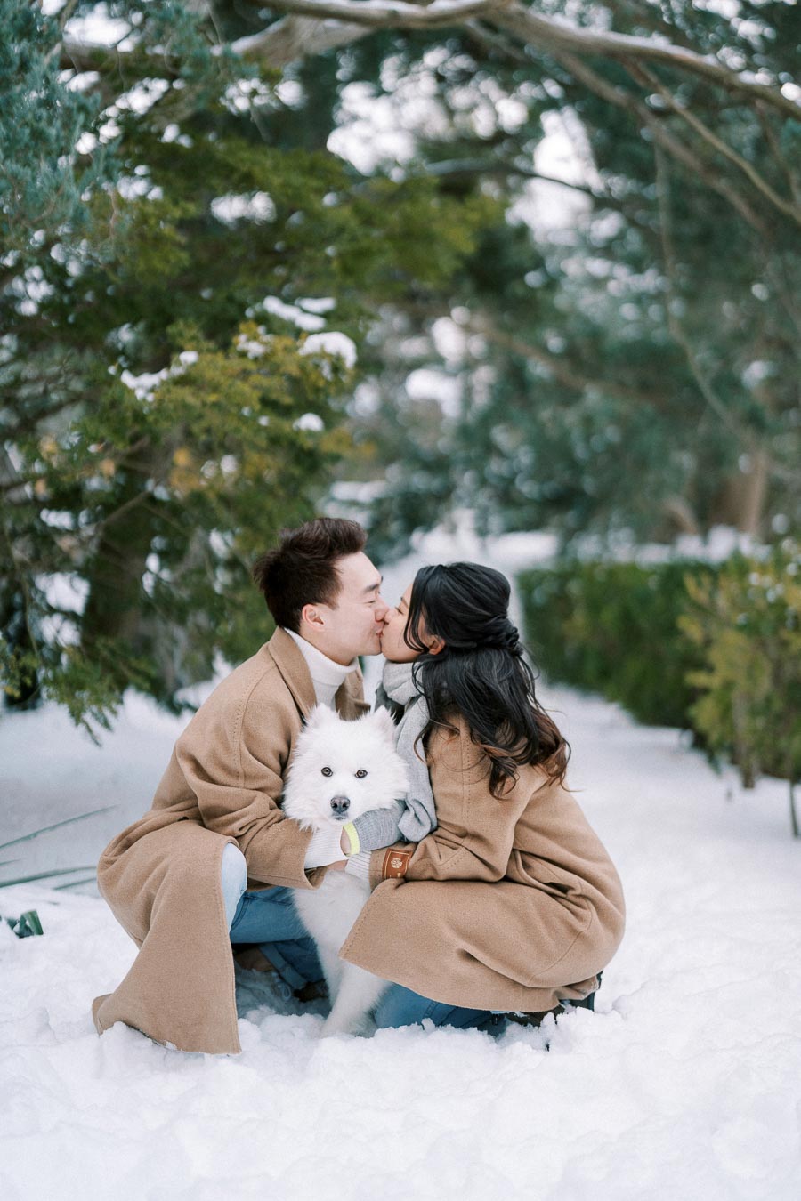 A couple dressed in matching brown coats kissing in a snowy landscape, with a fluffy white dog sitting between them.